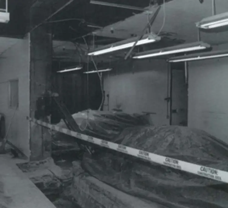 Black-and-white photo of an interior seismic retrofit work area with caution tape and excavated floor